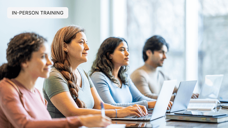 A group of four mature lenders sit together at a long desk, each with note books and laptops out in front of them as they take notes during a training.
